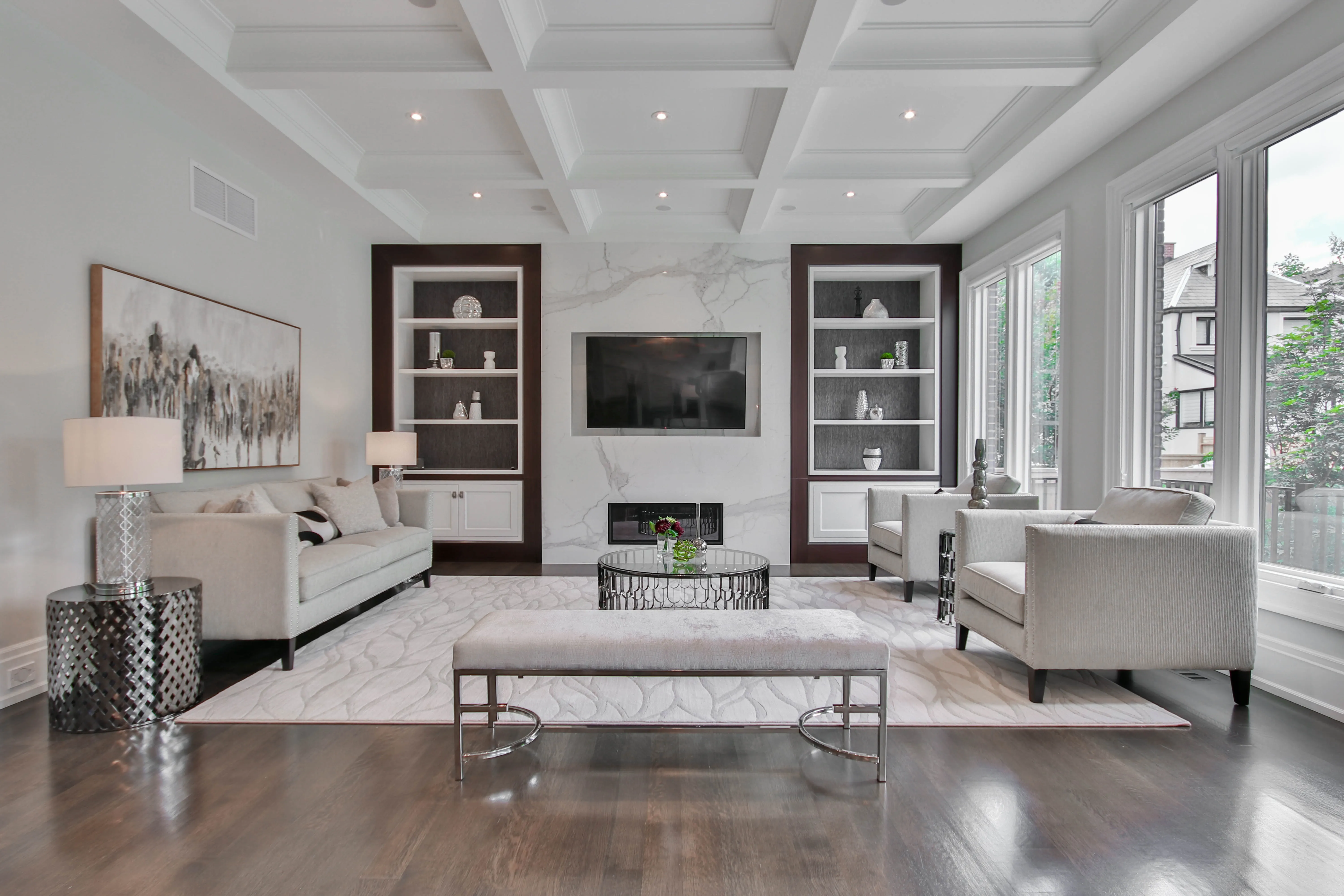 Bright modern living room with white furniture, large windows, built-in shelves, and a wall-mounted flat-screen TV over a fireplace.