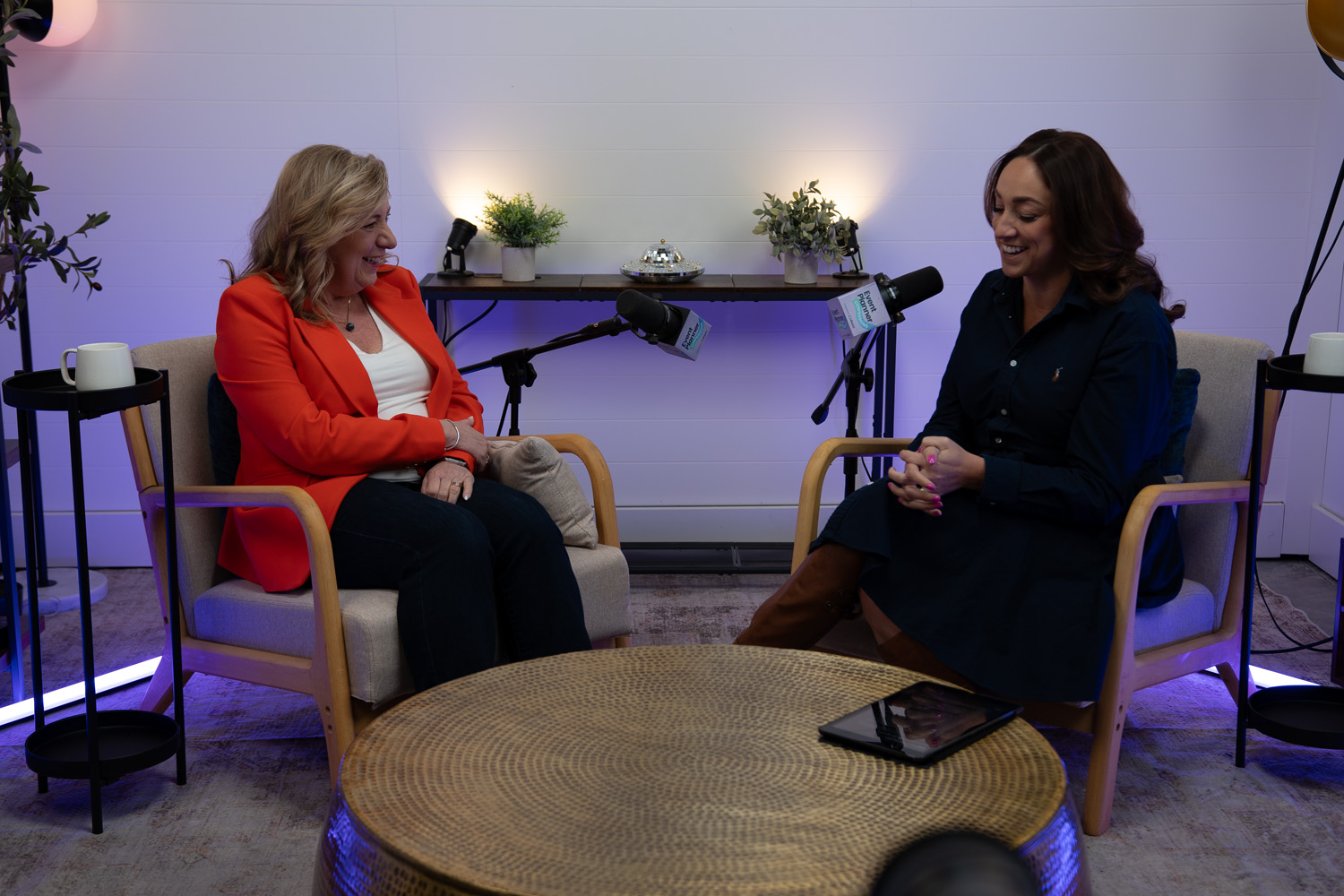 Two women seated in armchairs facing each other, smiling and talking in a cozy studio with microphones and soft purple lighting.