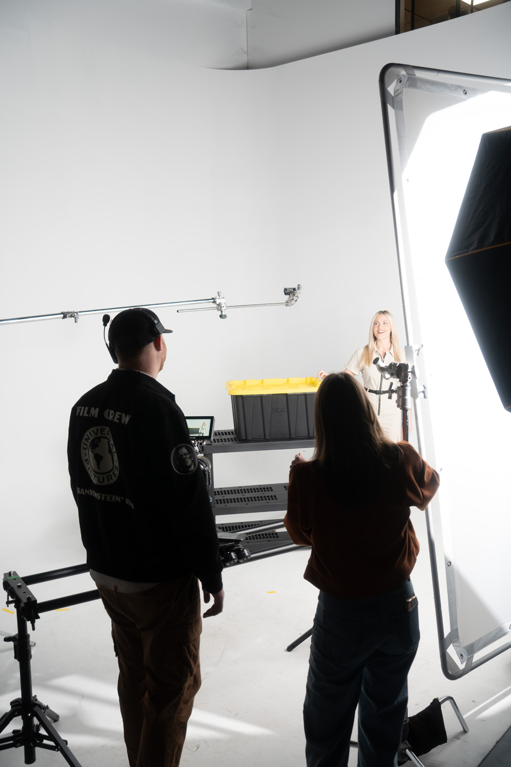 Two film crew members operating camera and lighting equipment filming a woman standing near a black storage bin with a yellow lid in a white studio.