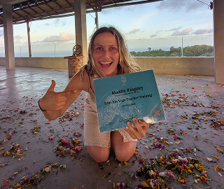 Maddie smiling and giving a thumbs up while holding her yoga teacher training certificate, kneeling on a floor scattered with flower petals