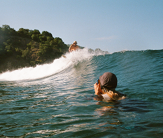 Film photo taken from the water of a surfer riding a breaking wave past a green headland, with another swimmer photographing in the foreground