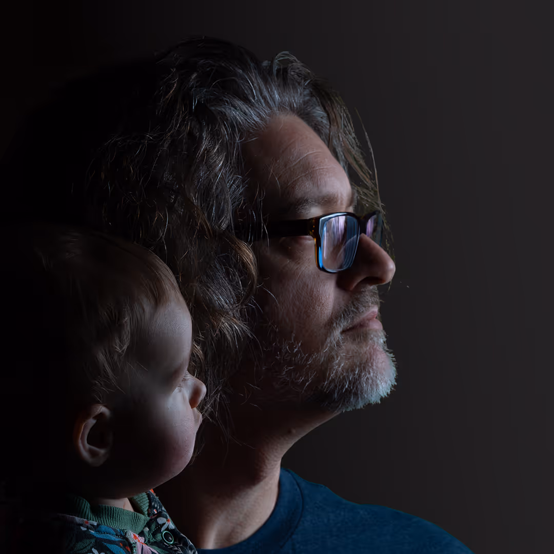 Side profile of a man with glasses and gray hair looking ahead, holding a baby also looking forward against a dark background.