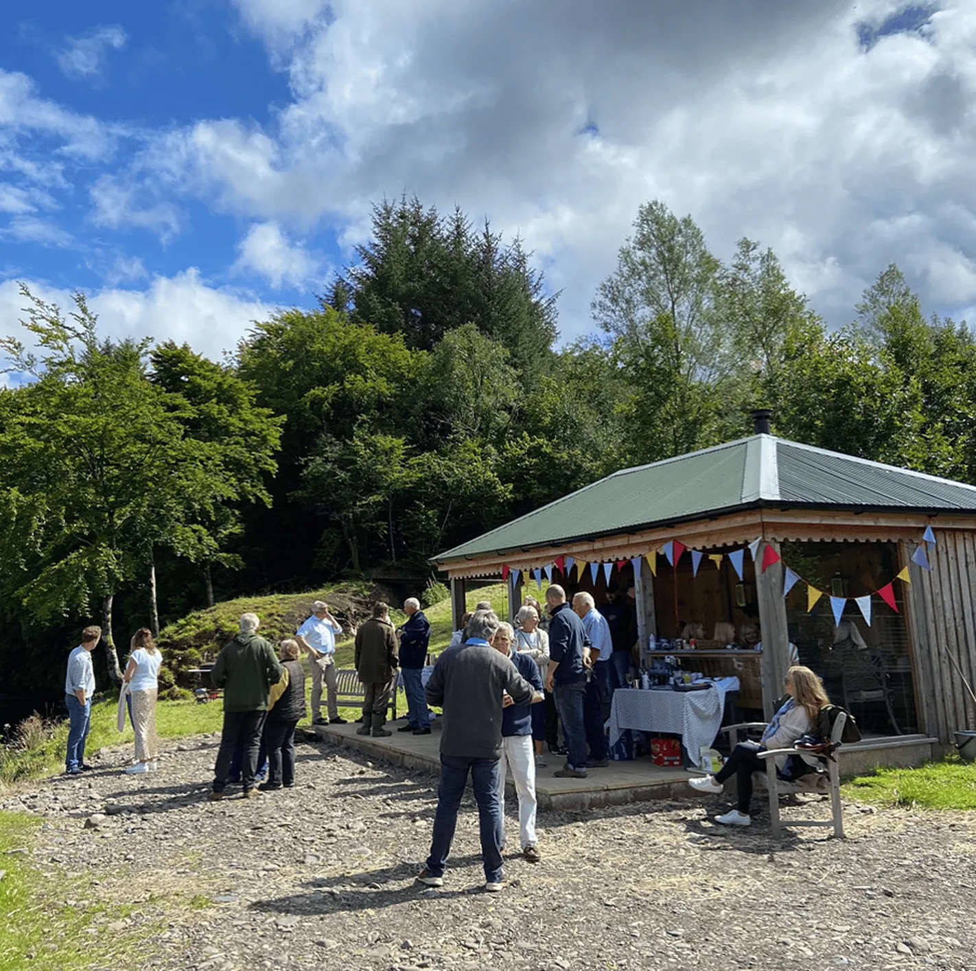 Group of people socialising outside a pavilion with colourful bunting, surrounded by trees under a partly cloudy blue sky.