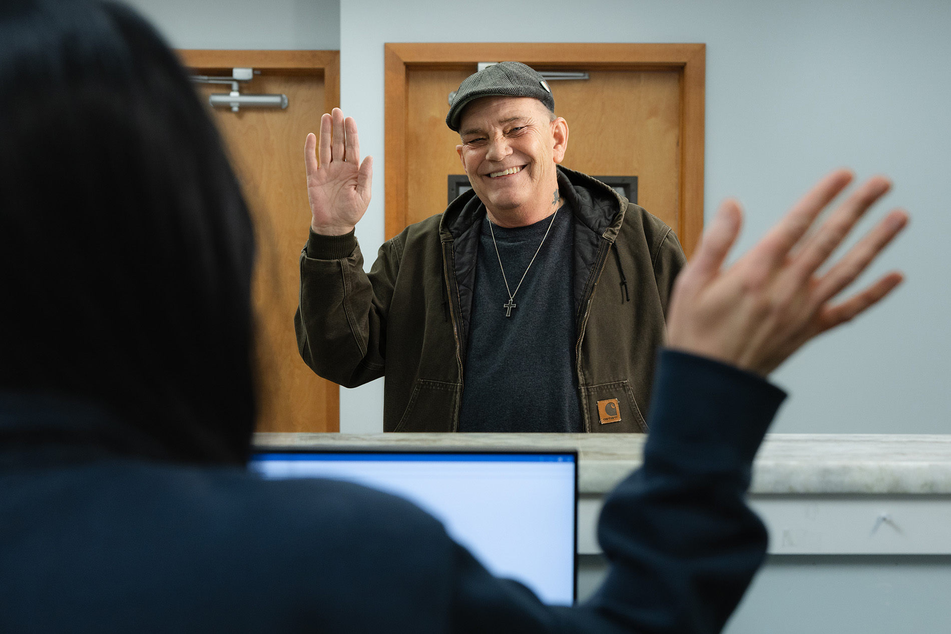 Smiling man in a jacket and flat cap raising his hand in greeting to a person behind a reception desk with a computer.