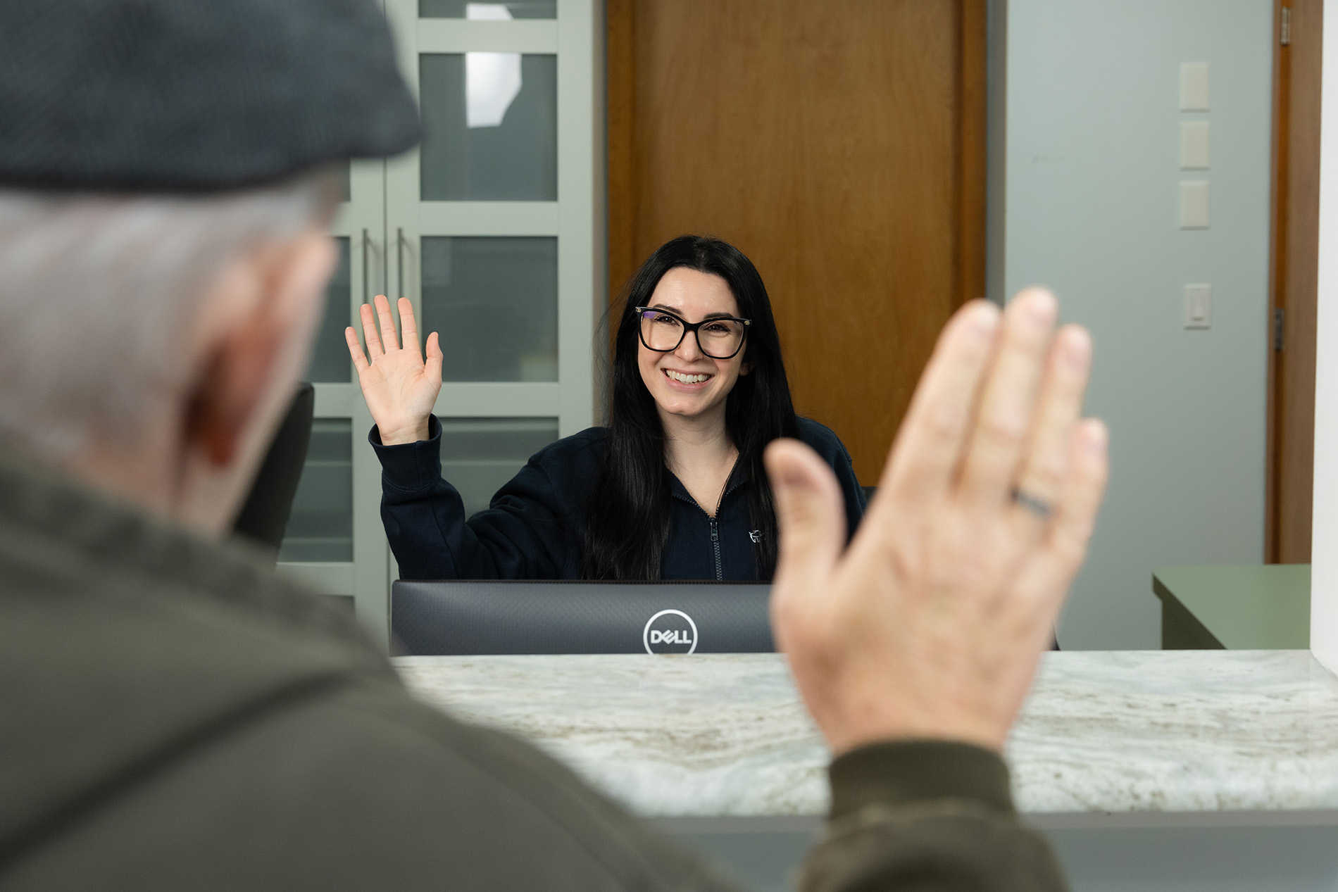 Smiling woman with glasses waving from behind a reception desk to an older man raising his hand in return.