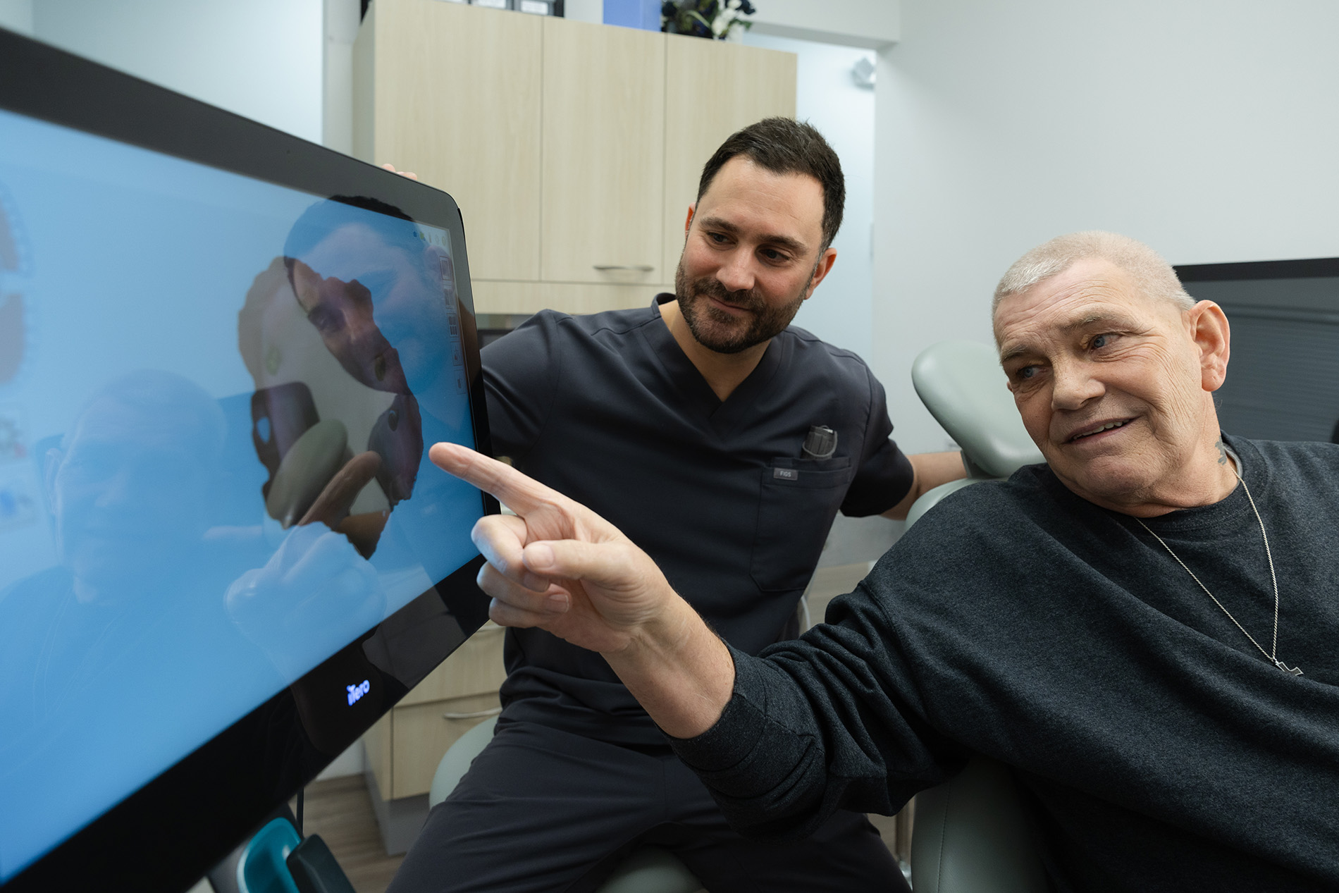 A male dental professional and an older male patient look at and point to a dental imaging screen in a dental office.