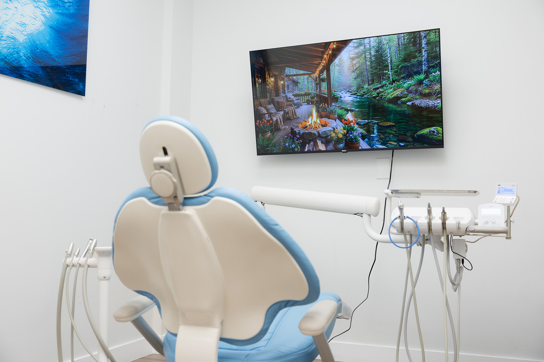 Dental examination room with a blue and white dental chair facing a wall-mounted TV showing a serene outdoor scene with a creek and a cabin porch with a fire pit.