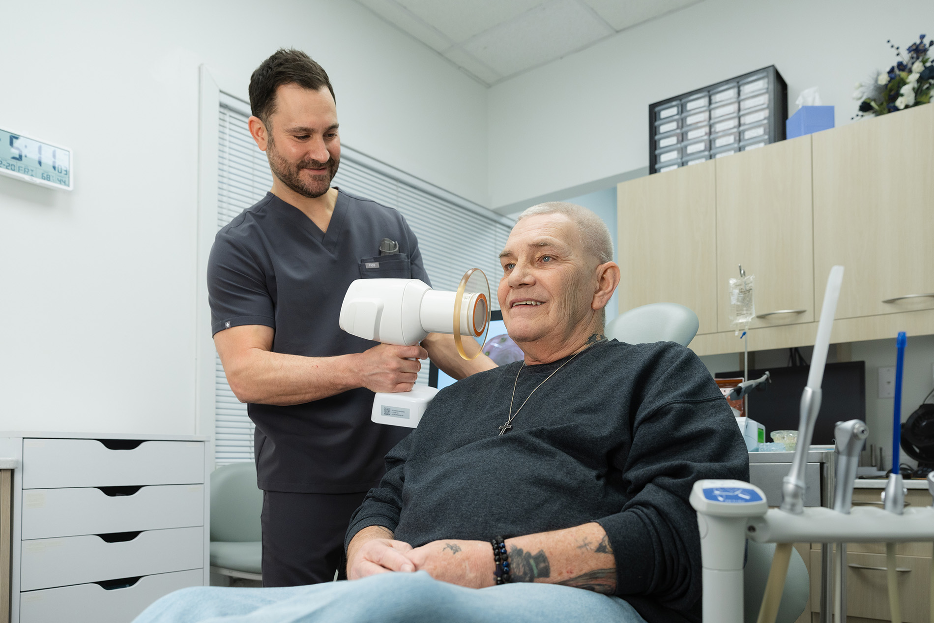 Dental professional using a handheld X-ray device on a seated older male patient in a dental office.