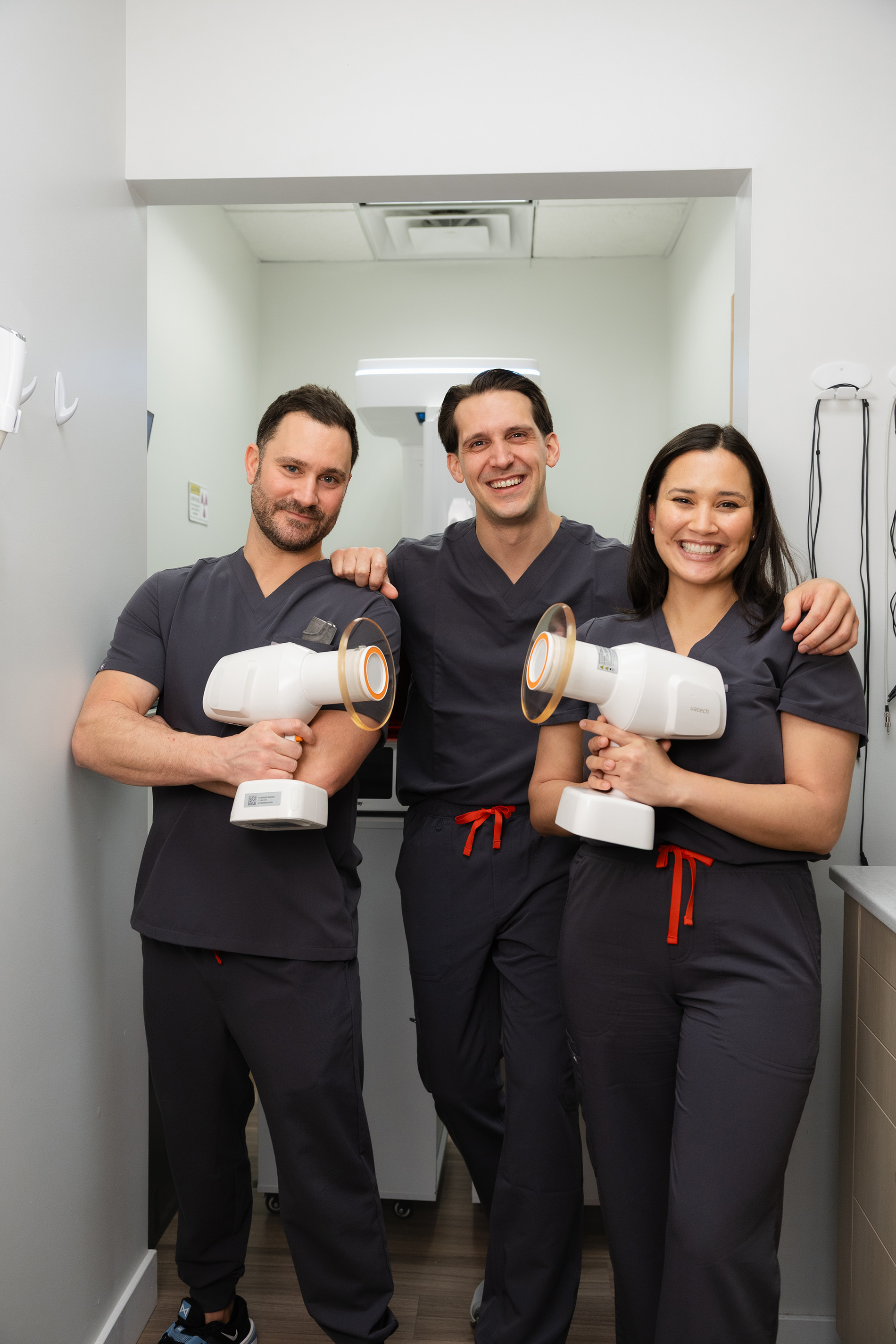 Three dental professionals in dark scrubs smiling and holding handheld X-ray devices in a clinic room.