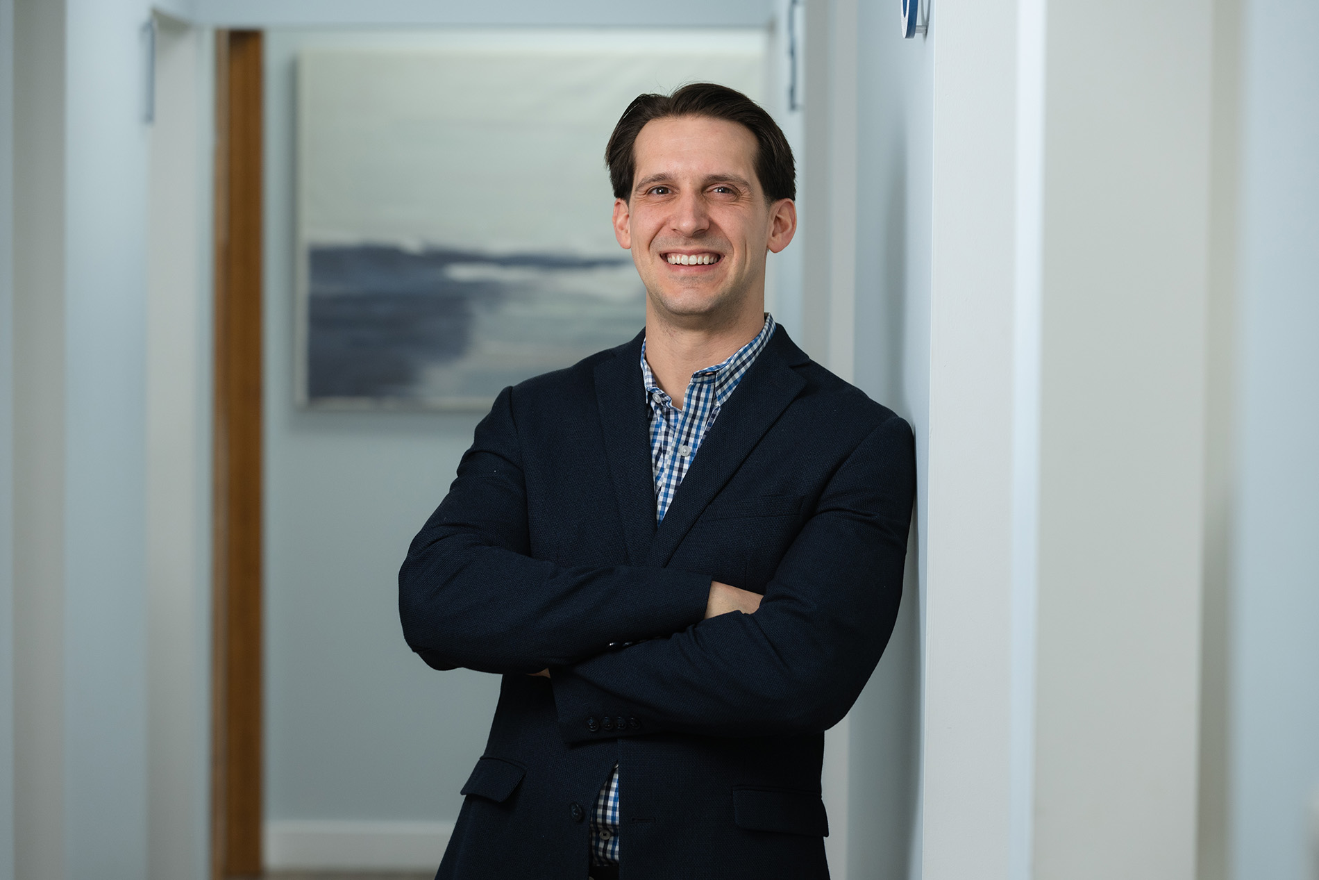 Smiling man in a dark blazer and checkered shirt standing with arms crossed leaning against a white wall in a hallway.