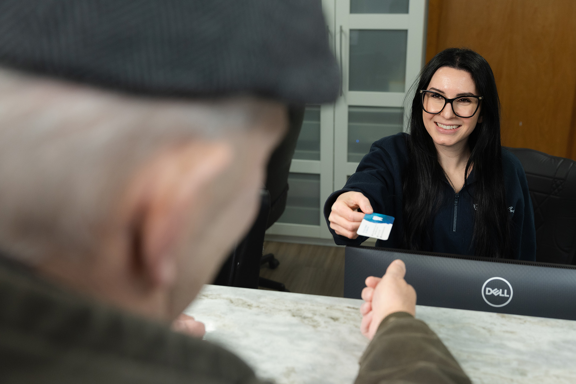 Smiling receptionist with black hair and glasses handing a card to an elderly man across a counter.