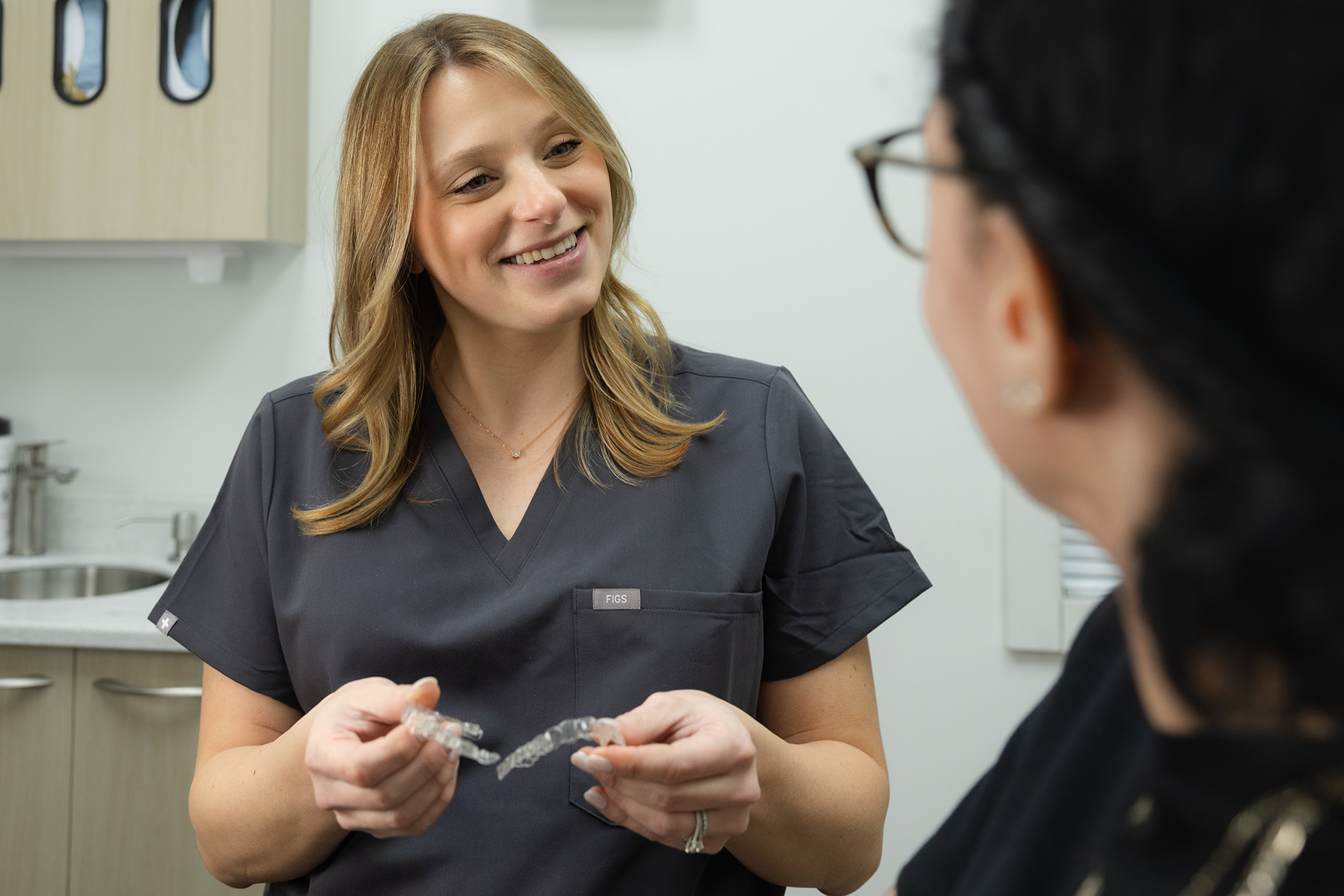 Smiling dental professional in gray scrubs holding clear dental aligners while talking to a patient.