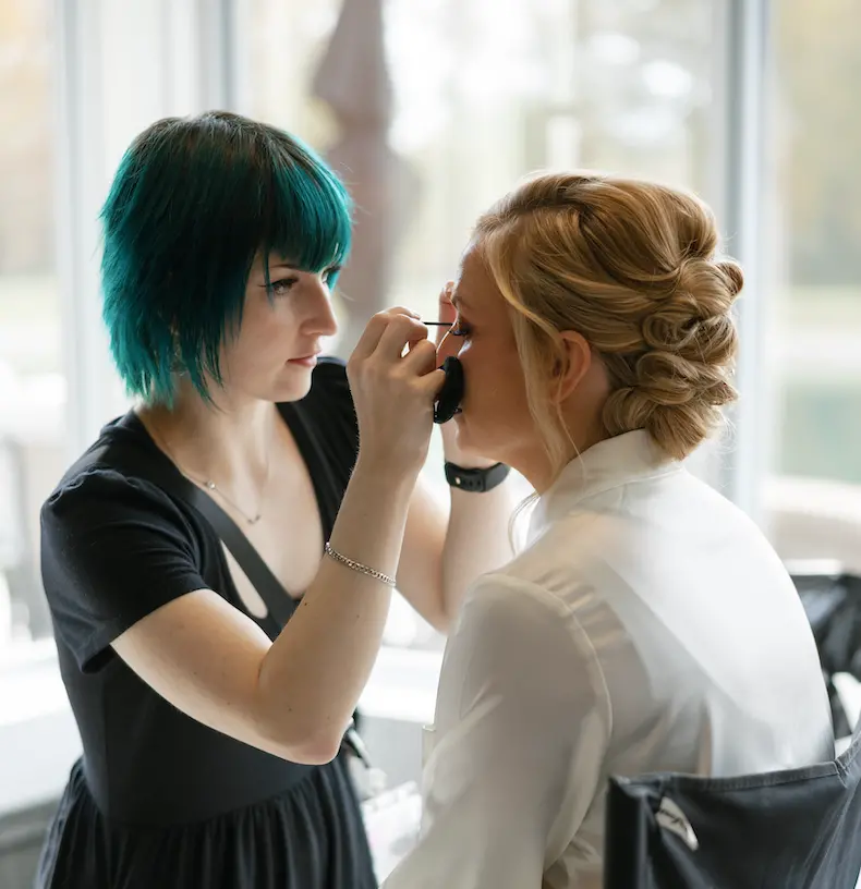 Our makeup artist Katie, doing makeup on a beautiful blonde bride that is wearing an updo. Katie has Blue short hair and a black dress on.