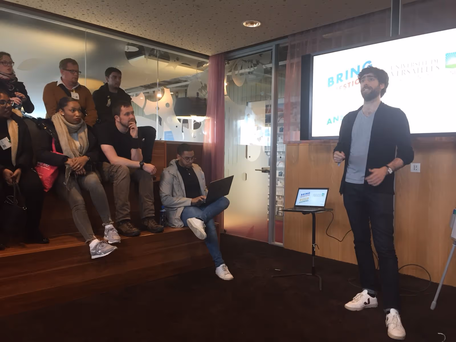 Hugo donnant une présentation devant un groupe assis sur des marches en bois, avec un ordinateur portable sur une petite table et un écran affichant du texte derrière lui.