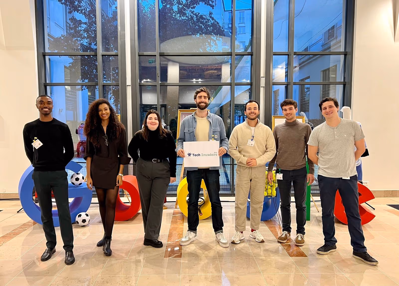 Sept personnes debout à l'intérieur devant de grandes fenêtres avec des lettres Google colorées et des ballons de football en arrière-plan, une personne tenant une pancarte Tech Invaders.