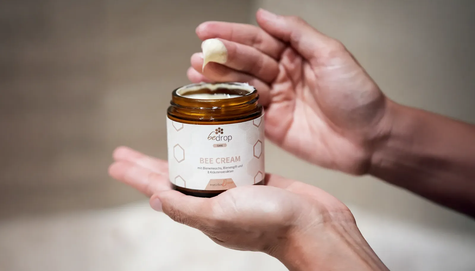  A person holding an open jar of "Bedrop Bee Cream" and scooping a small amount with their finger. The cream contains beeswax, bee venom, and herbal extracts, as shown on the label.