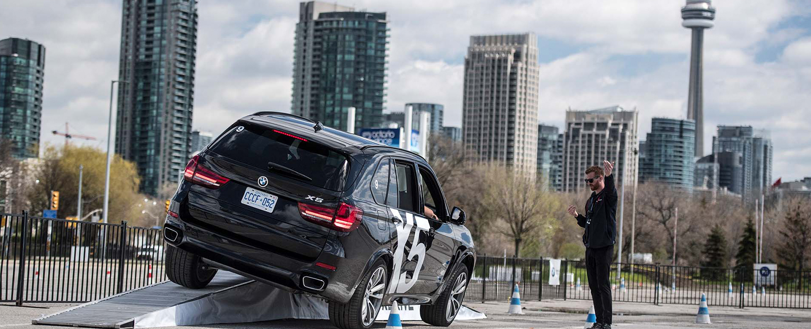 SUV driving up an angled ramp during a controlled driving exercise, with city buildings in the background and traffic cones marking the course.