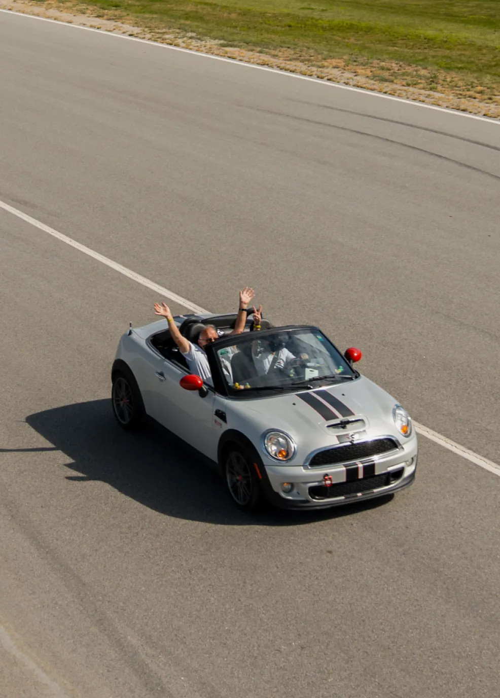 A guest in a silver MINI Cooper Convertible raises their hands in celebration while driving down a professional asphalt track