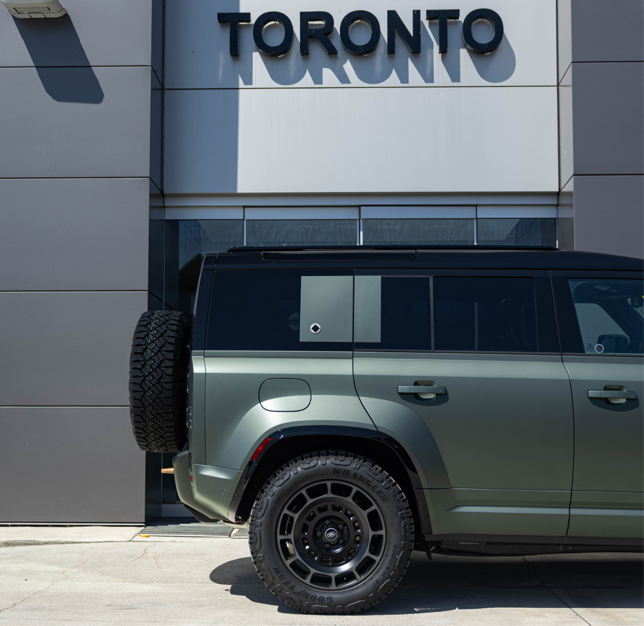 Side view of green Land Rover Defender parked at JLR Toronto dealership, highlighting rugged design and brand presence in urban setting.