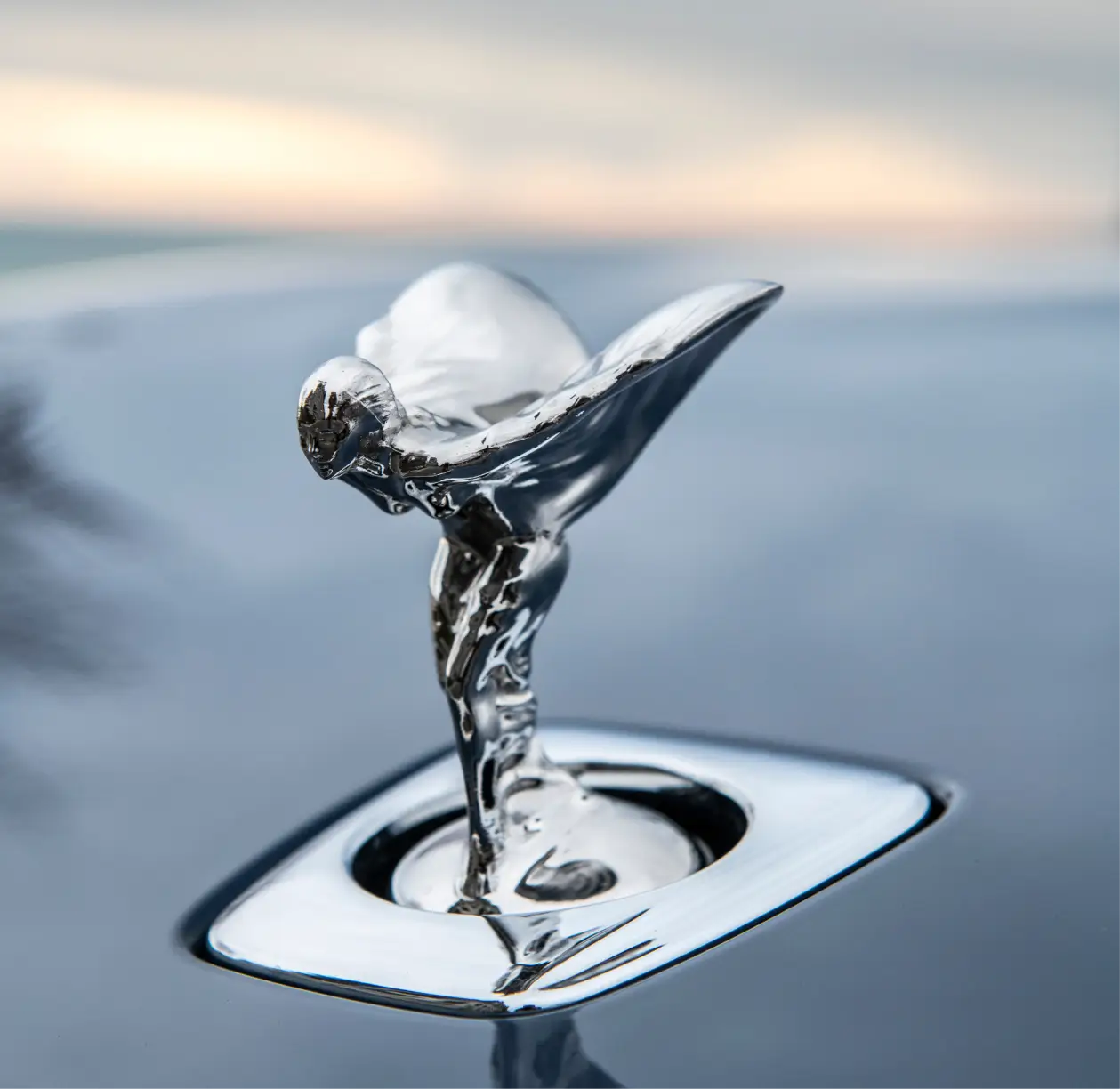 Close‑up of the Spirit of Ecstasy hood ornament reflecting sunset light, representing timeless craftsmanship and the artistry behind Rolls‑Royce design.