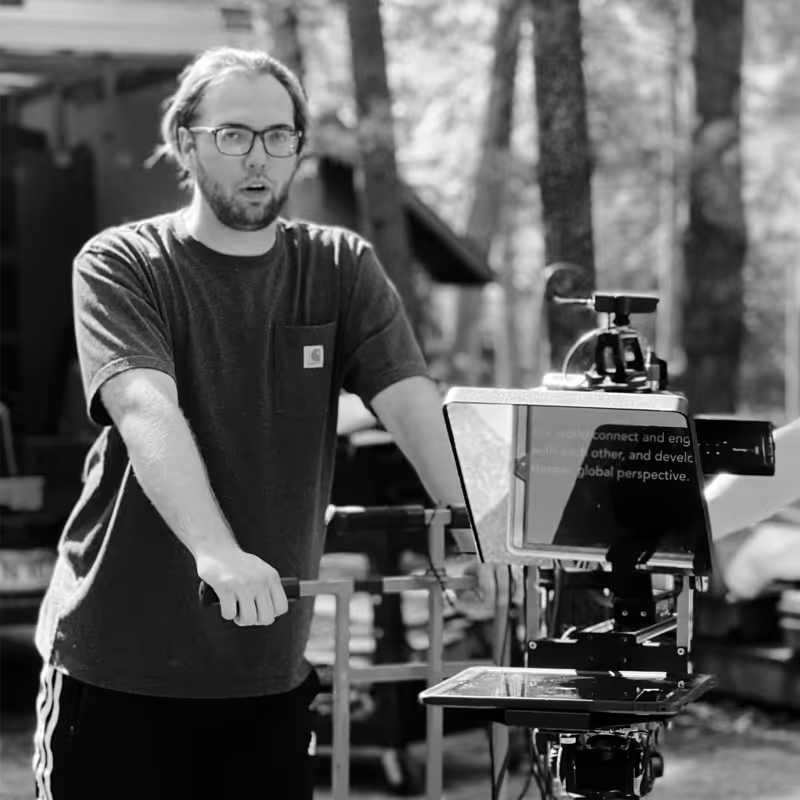 Man with glasses operating a teleprompter outdoors in a wooded area.