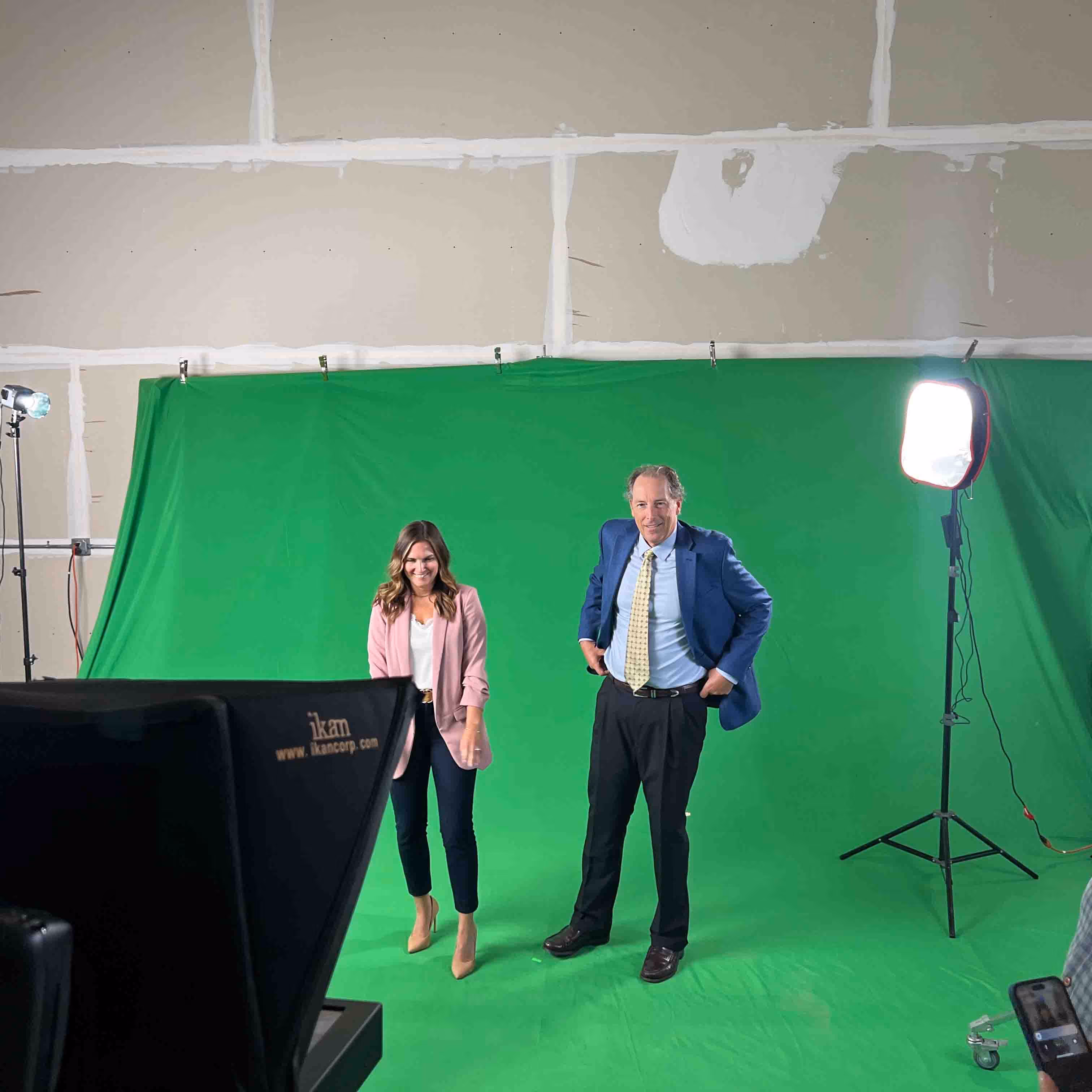 Two people standing in front of a green screen in a studio with professional lighting and camera equipment visible.