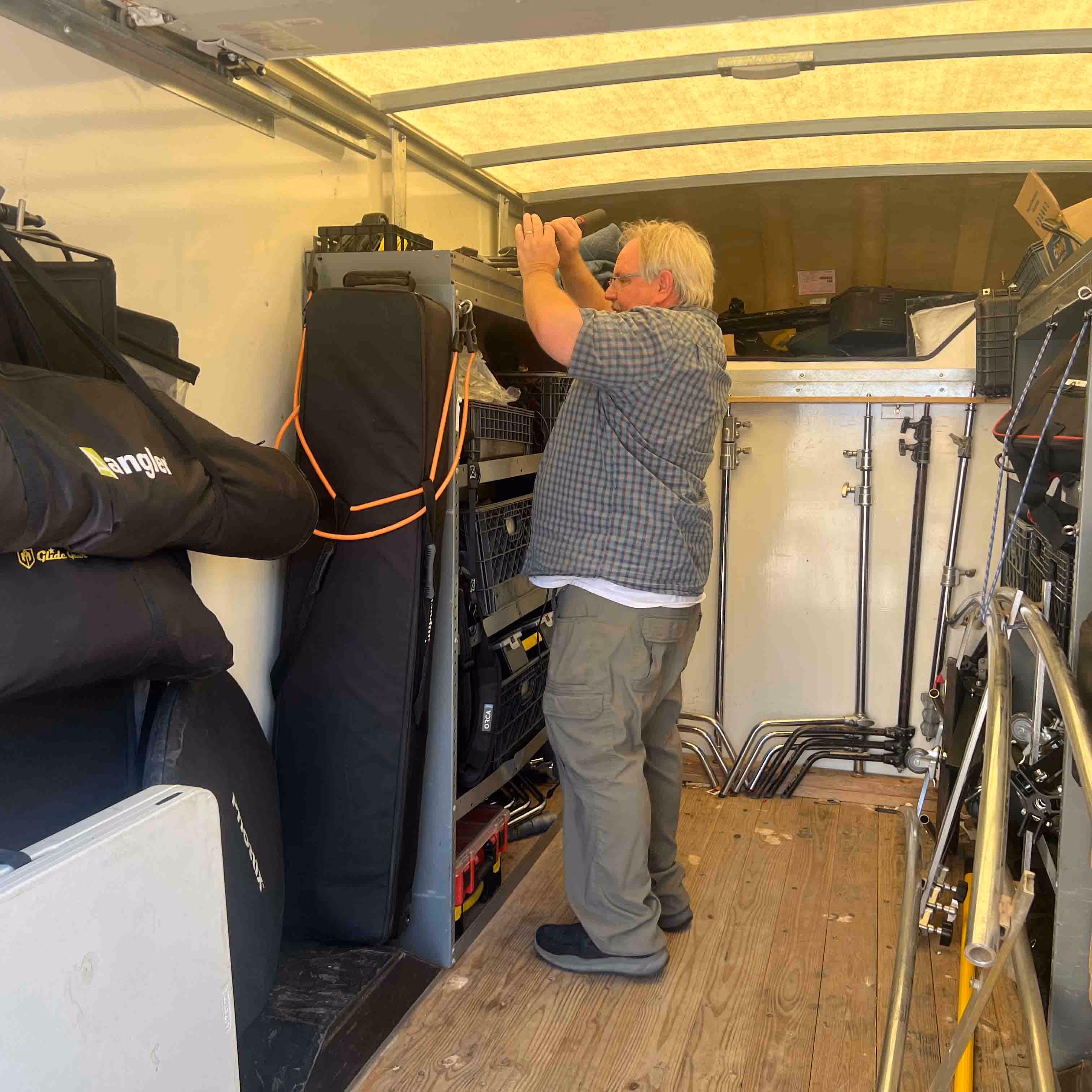 Man organizing or adjusting storage equipment inside a trailer filled with audio or filming gear.