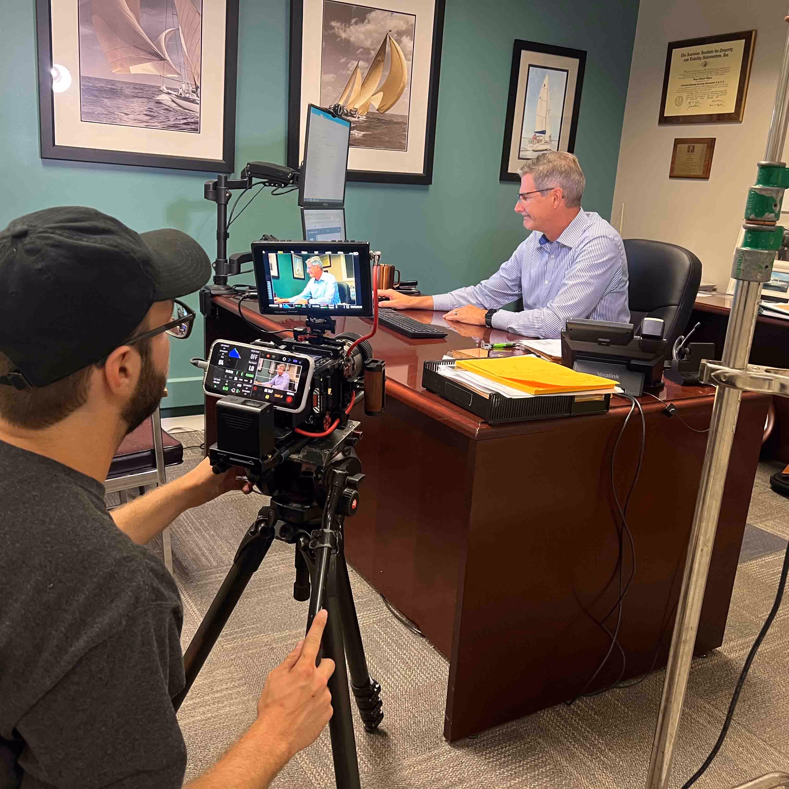 Man operating a professional camera on a tripod filming another man working at a desk in an office with framed sailboat pictures on the wall.