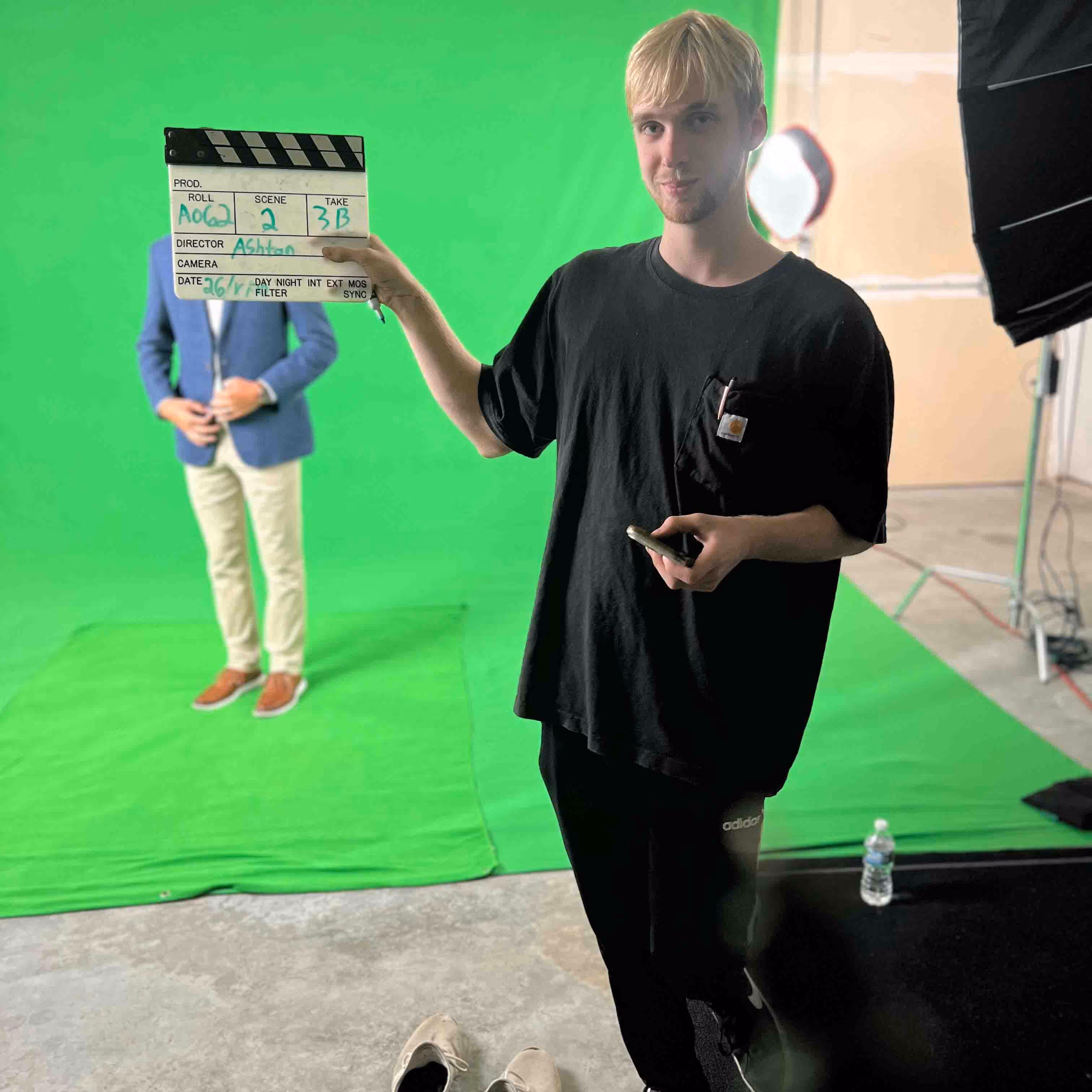 Man in black holding a film clapperboard on a green screen set with another person in a blue blazer in the background.