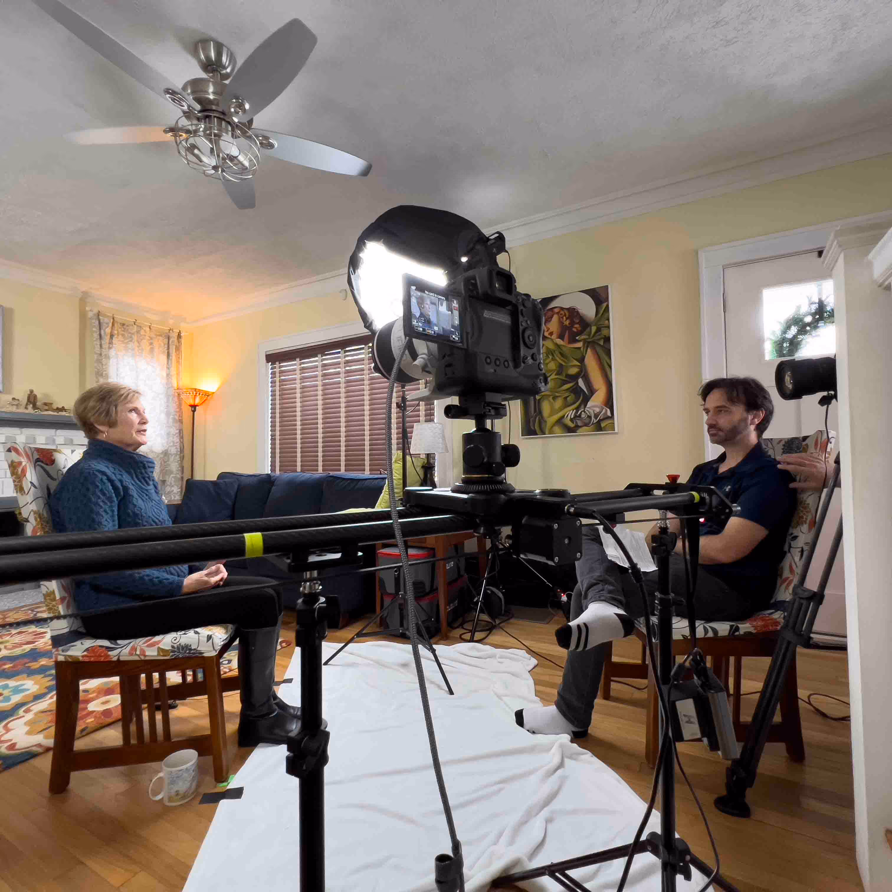 Two people seated on floral chairs facing each other in a living room set up for video recording with a camera, lighting, and a white backdrop on the floor.