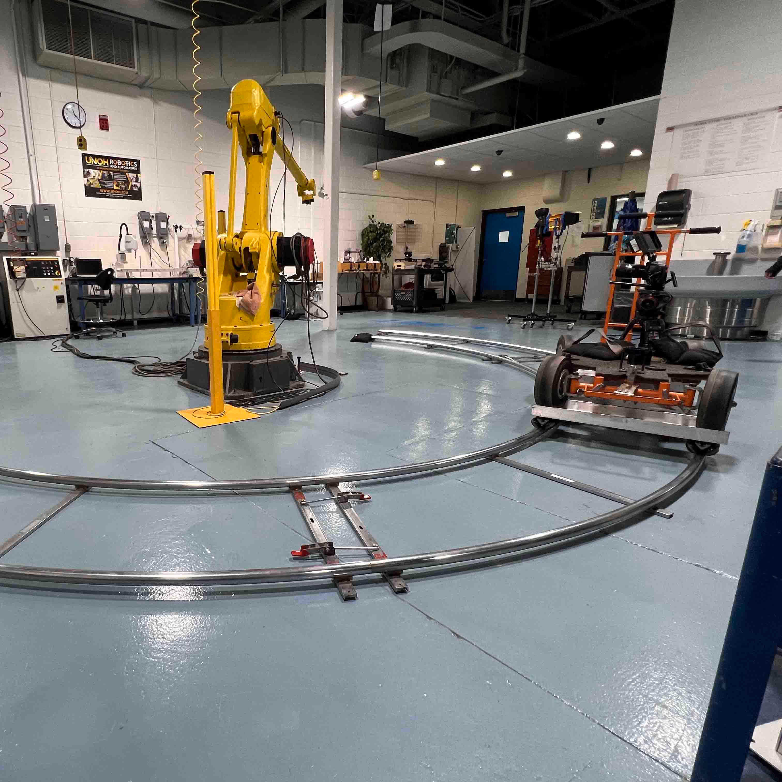 Yellow industrial robotic arm inside a workshop with a curved metal track and a wheeled camera dolly on the track.