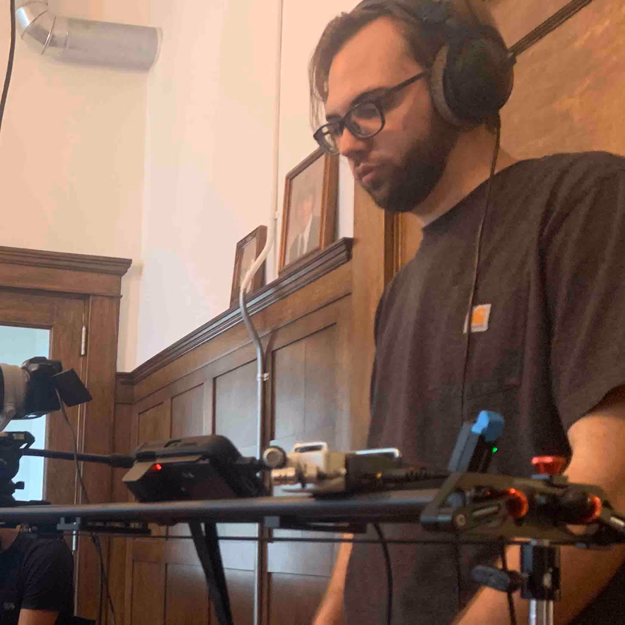 Man wearing headphones and glasses operating camera equipment on a tripod in a wood-paneled room.