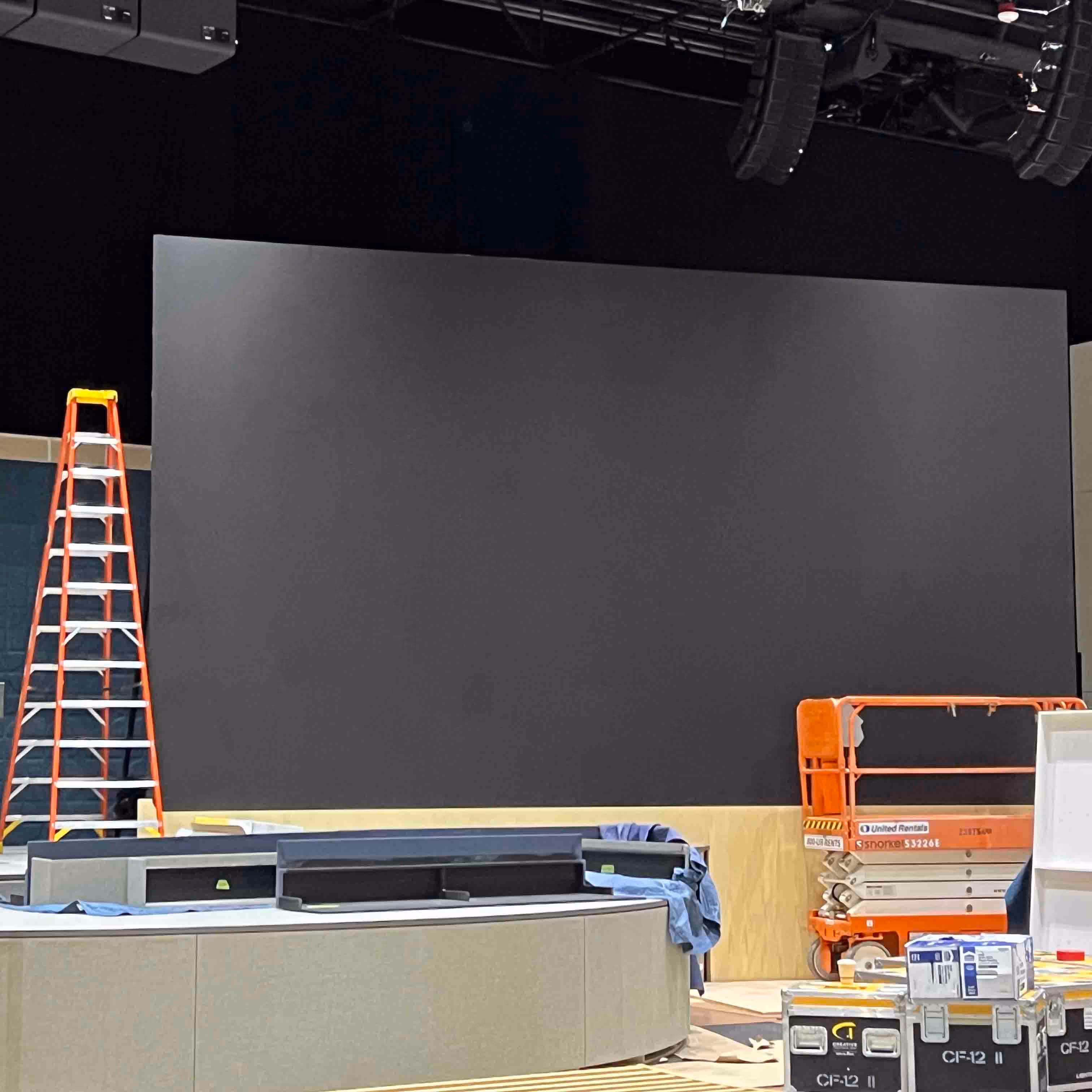 Indoor construction area with an empty black wall, a tall red ladder, an orange scissor lift, and various equipment cases and materials.