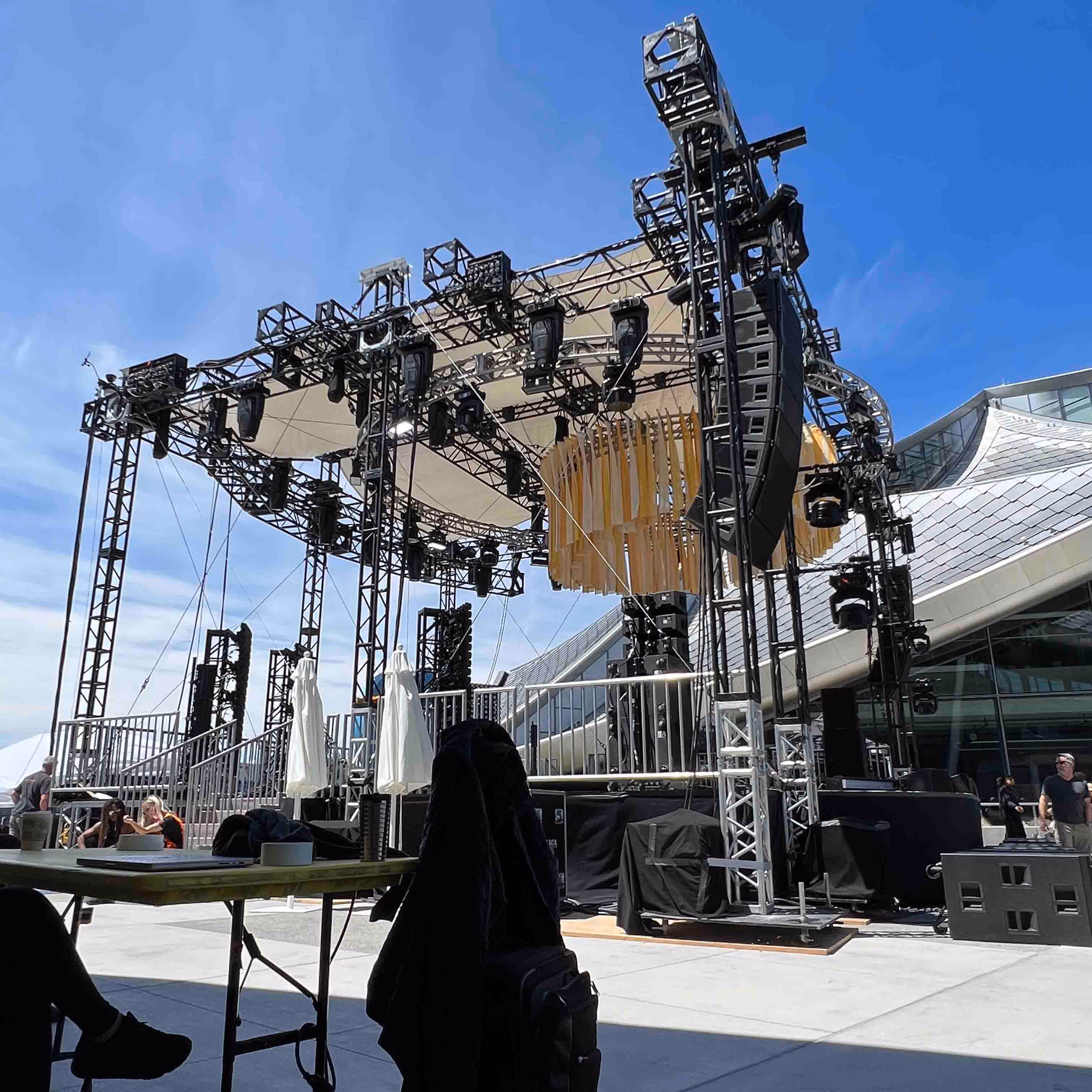 Outdoor stage setup with lighting, speakers, and a decorative wooden installation under clear blue sky near modern architectural buildings.