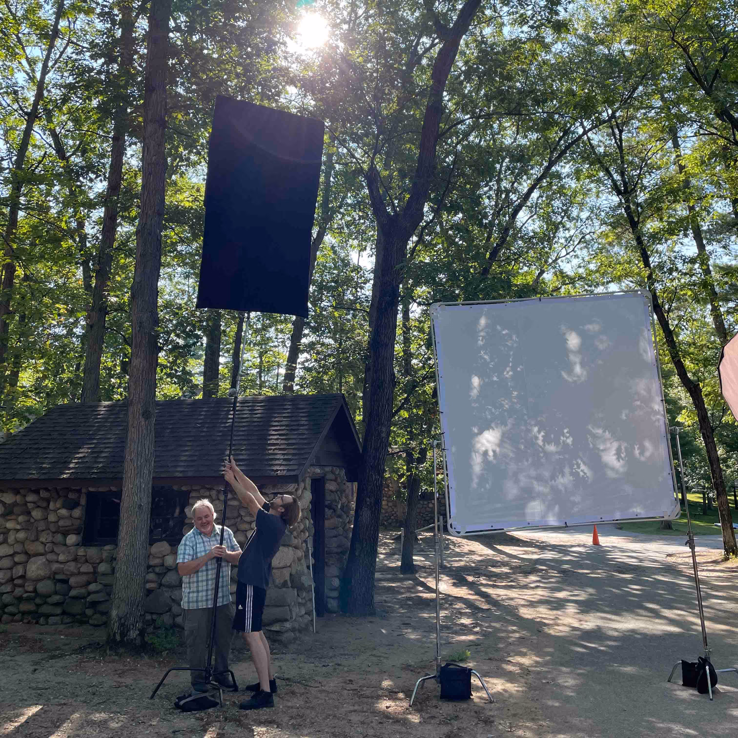 Two men adjusting a tall black fabric on a pole outdoors near a stone cabin surrounded by trees and sunlight.