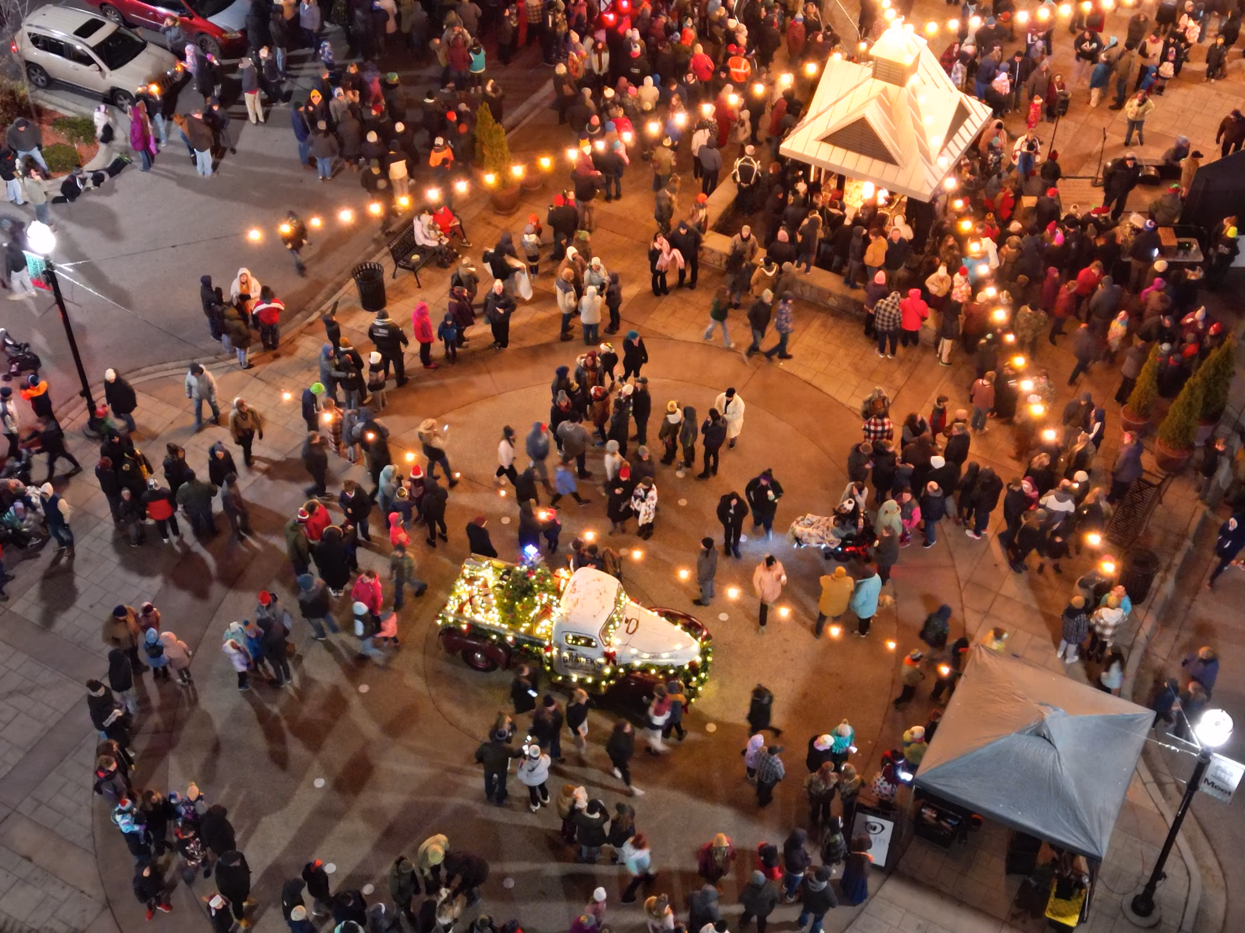 A nighttime aerial view of a festive crowd gathered around a small decorated truck adorned with Christmas lights in a public square.