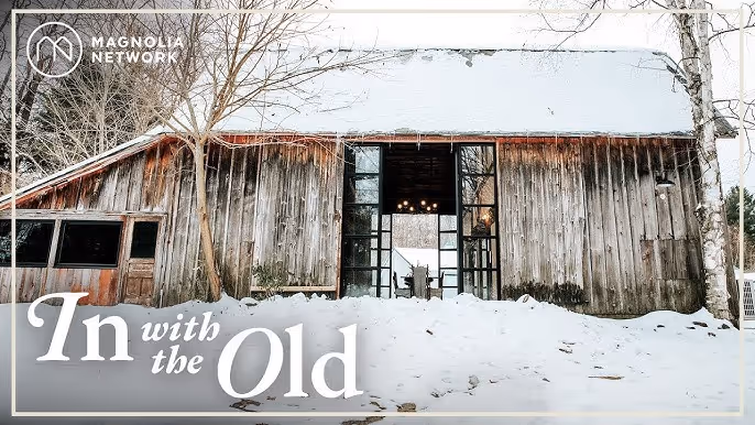 Rustic weathered barn with snow on the ground and bare trees in winter.