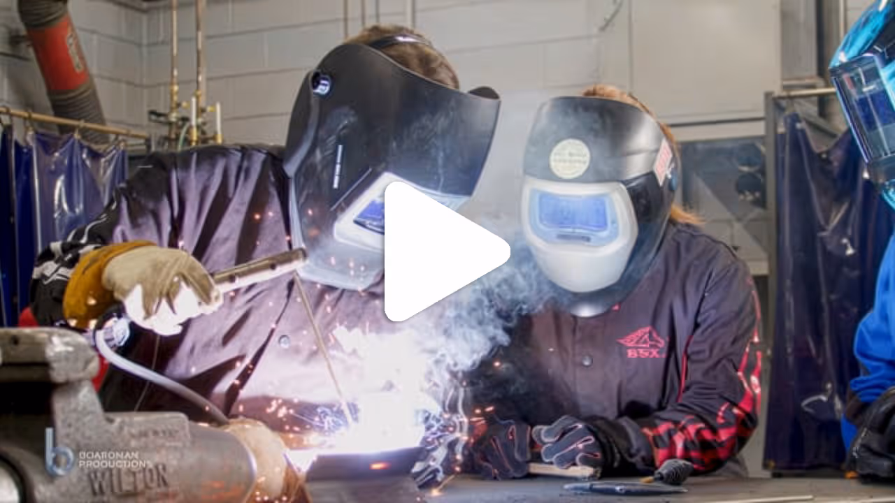 Two people wearing welding helmets and protective gloves working together on a welding project in a workshop.