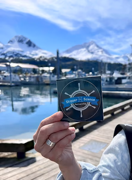 Hand holding a Skipper Science Partnership sticker with a ship's wheel design, by a marina with docked boats and snow-covered mountains in the background under a partly cloudy blue sky.