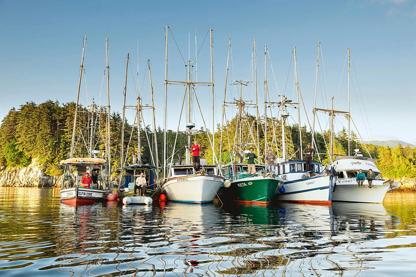 Seven sailboats docked side by side on calm water with six people standing or sitting on the boats, trees and rocky shore in the background under clear blue sky.