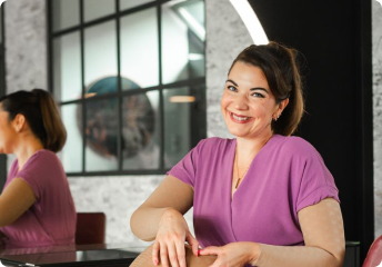 Smiling woman in a purple dress sitting cross-legged on a salon chair in front of a circular illuminated mirror.