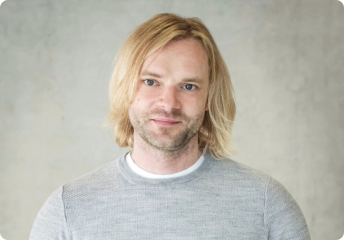 Smiling man with shoulder-length blonde hair wearing a light gray sweater against a neutral background.