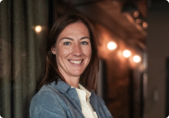 Smiling woman with brown hair wearing a light-colored shirt and gray jacket standing indoors with blurred warm lights in the background.
