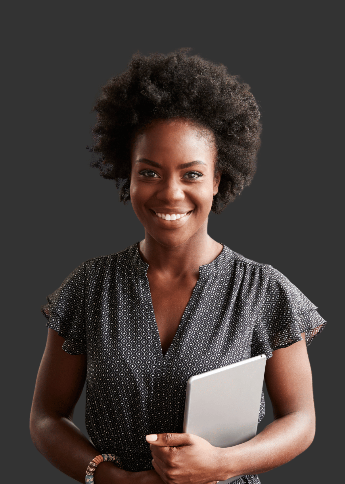 Smiling woman with natural curly hair wearing a patterned short-sleeve blouse, holding a tablet against a dark background.
