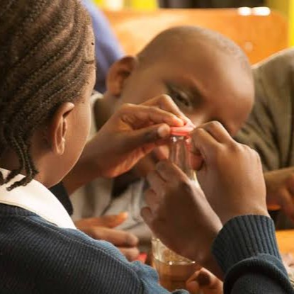 Two children, one with braided hair and the other leaning in closely, engage in an activity involving a small jar with a reddish lid, focusing intently on opening or closing it.