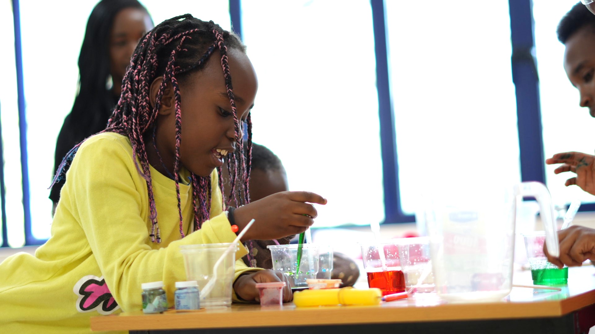 A young girl with pink and black braided hair wearing a yellow sweater is stirring a green liquid in a plastic cup at a table with various other colored liquids and small containers.