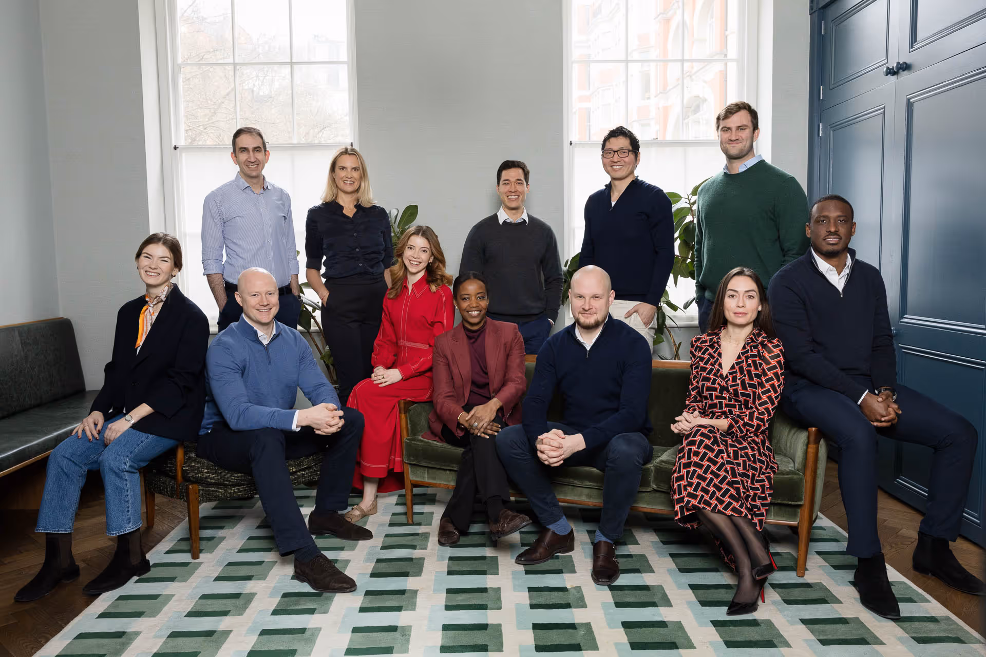 Group portrait of nine diverse professionals smiling, with four sitting on chairs and a couch, and five standing behind them in a modern office setting.