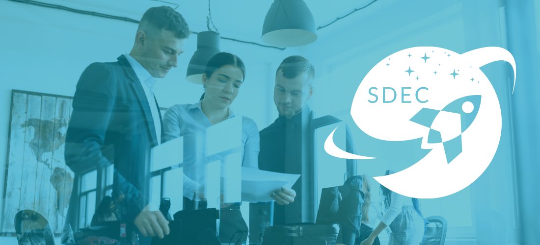 Three colleagues stand and look down at a piece of paper being held by the person in the center. To the left of the people is the SDEC logo. 