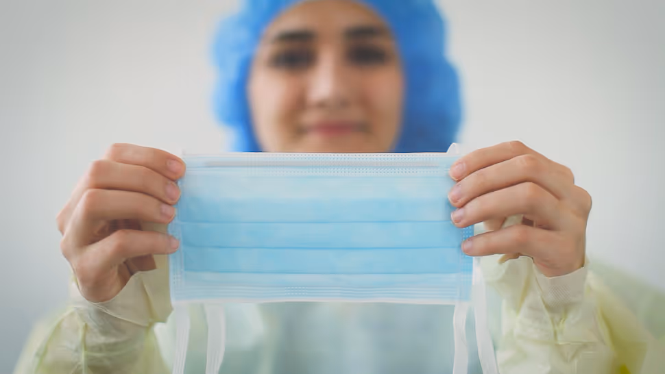 medical professional holding surgical tie mask up with the coloured side facing away from their face.