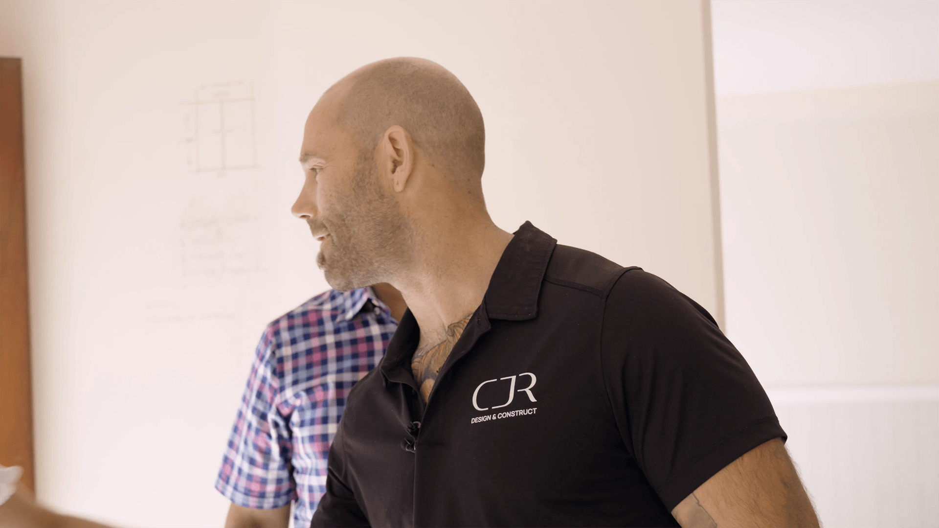 Man with a shaved head and beard wearing a black shirt with 'CJR Design & Construct' logo, looking to the left, with another man in a checkered shirt blurred in the background.